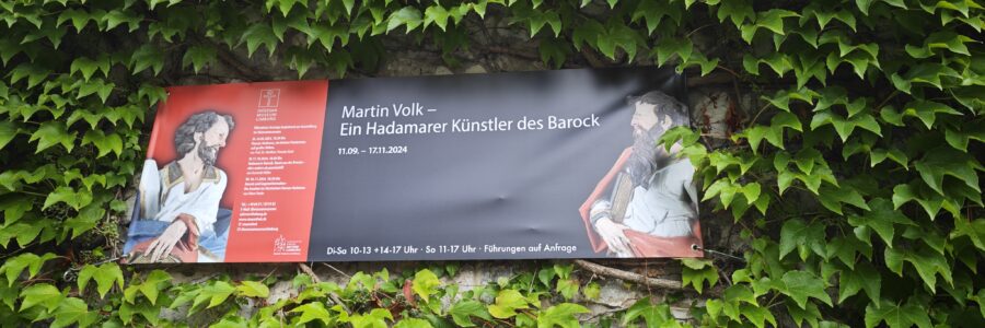 Auf dem Bild ist das Plakat zur Ausstellung von Martin Volk inmitten von grünen Pflanzen