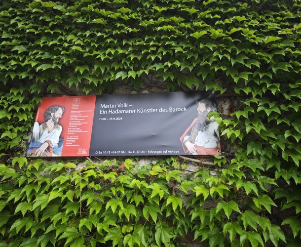 Auf dem Bild ist das Plakat zur Ausstellung von Martin Volk inmitten von grünen Pflanzen
