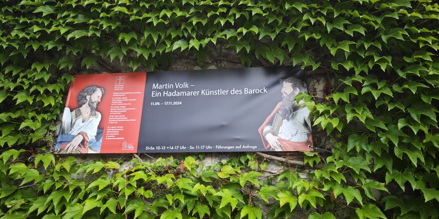 Auf dem Bild ist das Plakat zur Ausstellung von Martin Volk inmitten von grünen Pflanzen
