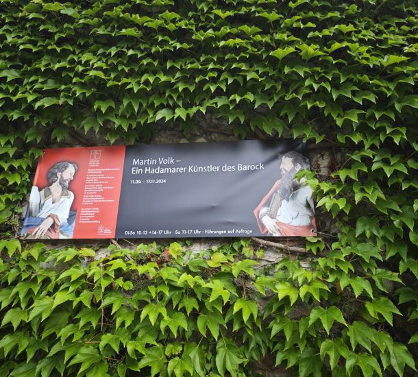 Auf dem Bild ist das Plakat zur Ausstellung von Martin Volk inmitten von grünen Pflanzen